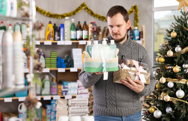 Selective middle-aged male customer choosing present in cosmetic department of retail store decorated for Christmas