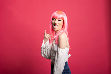 Trendy pink haired woman shows rock hand sign and sticks out her tongue while smiling. Confident...