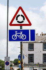 UK road signs for mini-roundabout and cycle route on a rusty post. An urban scene is blurred in the...