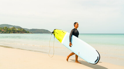 Confidence Asian man in sportswear enjoy water sports surfing at the sea on summer beach holiday vacation. Healthy people outdoor active lifestyle holding surfboard walking on the beach in sunny day.
