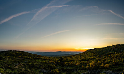 A wide mountainous landscape at sunset, with hills covered with vegetation under a clear blue sky...