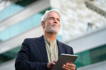 Senior businessman in suit using tablet and looking away thoughtfully outside office building. Professional and focused in urban environment