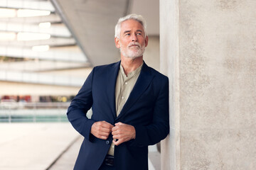 Senior businessman in formal outfit standing outdoors near concrete wall, buttoning blazer. Calm...