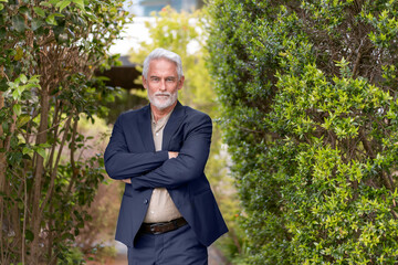 Senior businessman in formal suit standing with crossed arms in green outdoor setting. Calm and...