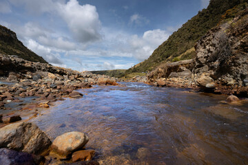 A vibrant, clear river meanders through a rocky valley, flanked by lush green mountains under a...