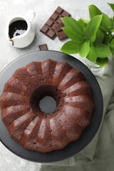 Tasty bundt cake, pieces of chocolate, sauce and green leaves on grey table, flat lay