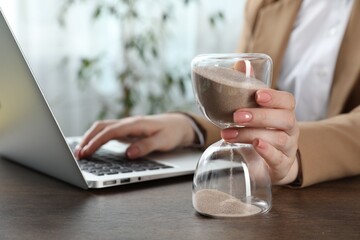 Deadline. Woman with hourglass and laptop working at wooden table in office, closeup