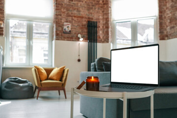 Laptop with blank screen and burning candle on a small round table in bright loft interior with...