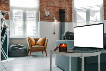 Laptop with blank screen and burning candle on a small round table in bright loft interior with...