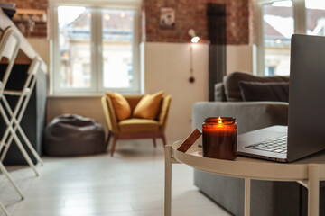 Burning candle and laptop on small round table in cozy loft interior with mustard armchair, bean...