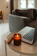 Burning candle and laptop on small round table in cozy loft interior with mustard armchair, bean bag, gray sofa, and bright windows. Warm and relaxed home workspace