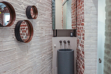 Stylish industrial bathroom corner with red brick walls, white textured surface, round mirrors,...