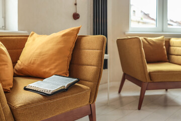 Cozy mustard yellow armchair with soft cushions and an open Bible placed on the seat, near a white side table and radiator, in a bright, peaceful corner