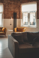 Obraz premium Bright loft-style living room with gray sofa, mustard armchairs, laptop, white table, brick walls and large windows. Cozy and modern interior workspace