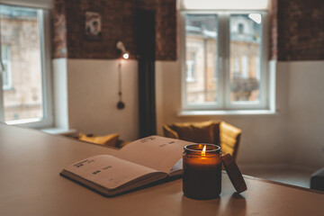 Burning candle and open planner on a table in cozy loft interior with warm light, mustard chairs,...