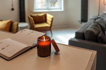 Burning candle and open planner on a table in cozy loft interior with warm light, mustard chairs,...