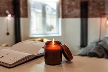 Burning candle and open planner on a table in cozy loft interior with warm light, mustard chairs,...