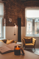 Burning candle in glass jar and open planner on table with soft background of cozy loft interior, mustard armchairs, brick wall, and daylight from large windows