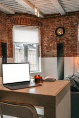 Laptop, burning candle and open planner on table in a bright loft-style living room with brick walls, mustard armchair and large windows letting in daylight