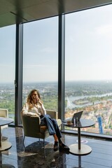 A sophisticated businesswoman relaxes in her elegant office, enjoying a panoramic view of the vibrant city skyline and calm river, reflecting professionalism, success, and modernity