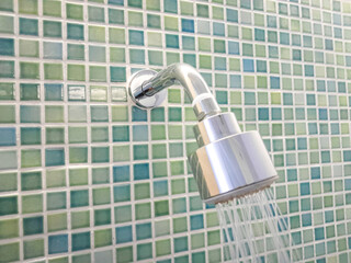 Modern Shower Head with Running Water Against Mosaic Tile Wall. A sleek metal shower head releasing...