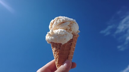 A hand holding an ice cream cone with a scoop of vanilla ice cream against a bright blue sky backdrop