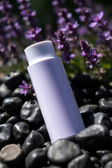 A minimalist white bottle rests on smooth black pebbles with a soft focus background of delicate purple flowers