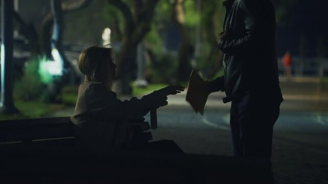 Shady handoff in a dark park &mdash; a woman on a bench discreetly passes an envelope to a standing man. Nighttime secret meeting with suspenseful, criminal or espionage undertones. secret agent word. night