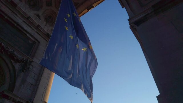European emblem sways slowly beneath grand stone arch against deep blue afternoon sky.