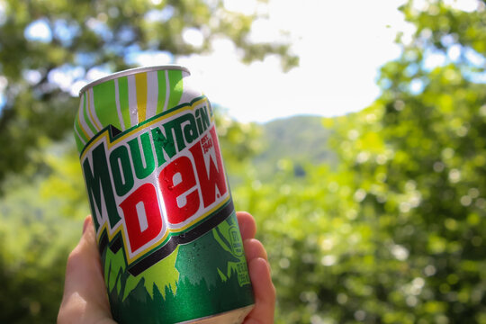 Close-up of a hand holding a can of Mountain Dew carbonated beverage outdoors with a mountain and light effect background.