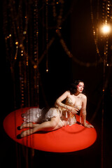 Young Caucasian woman in ornate golden corset, lying on red round stage floor, under hanging beads, dark background, front view. Concepts: cabaret, circus, retro, burlesque.
