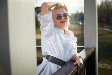 Woman in white dress and sunglasses poses confidently on a balcony during golden hour in a serene...