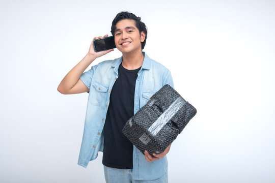 Smiling Southeast Asian delivery man holding a parcel while talking on phone, confirming delivery address. Isolated on a white background.