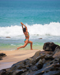Slim fit woman in a blue bikini doing yoga poses on the sandy rocky beach in Mirissa. Lunge forward and hands above the head.