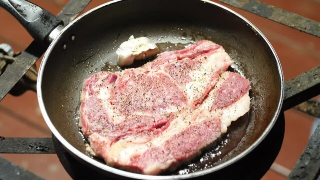 A close-up shot of a raw, seasoned ribeye steak being pan-seared. Garlic cloves are added, followed by a generous pat of butter, illustrating the process of cooking a delicious, juicy steak. 