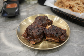 Grilled beef ribs are served on a golden plate, looking juicy and charred, accompanied by crispy fried enoki mushrooms and spicy sambal on the side.