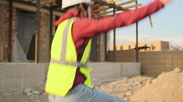 A construction worker in a hard hat and vest jumps to the music playing through his small earbuds, moving with wild freedom as the rhythm takes over and unleashes his pent up energy.