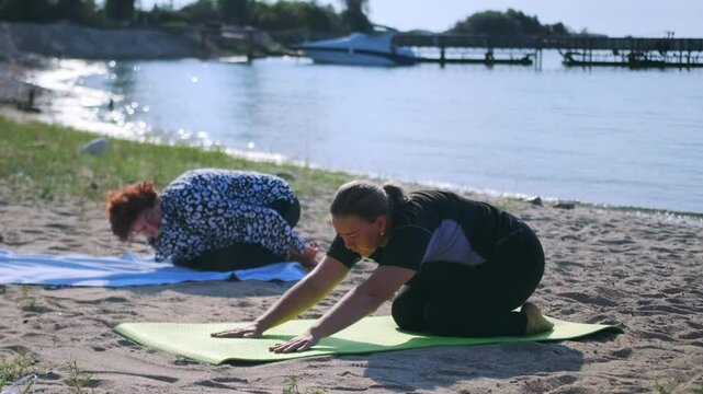 A mature woman and her adult daughter practice yoga by a scenic lake, a yacht and pier behind them. From a tucked position, they stretch forward, lengthening their spines and relaxing their bodies.