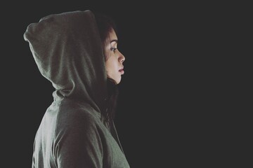 Thoughtful young woman in hooded shirt standing against black background