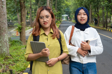 Two female students walking side by side on a university path, holding books and a tablet. Concept...