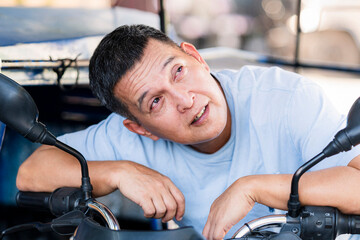 A Southeast Asian tricycle driver leans on his handlebars and gazes toward the sky, possibly...