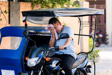 A tired Southeast Asian tricycle driver in casual wear wipes sweat from his face, suffering from...