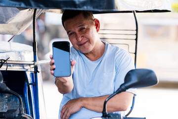 A cheerful Southeast Asian tricycle driver smiles warmly at the camera while proudly showing his...