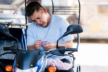 A worried Southeast Asian tricycle driver counts a small amount of money, looking down with...