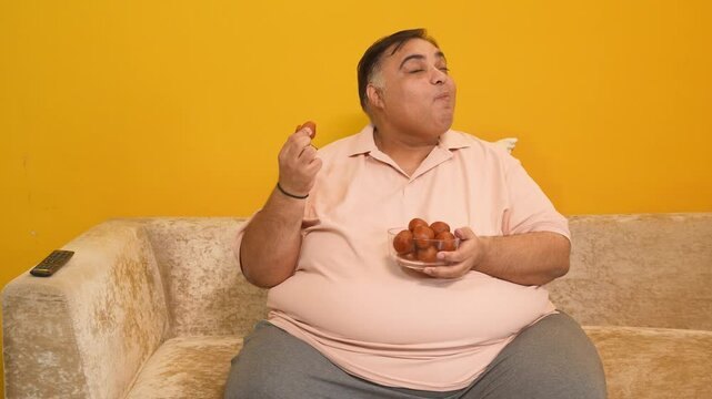 Overweight indian man eating gulab jamun sweets. Fat guy enjoy delicious food dessert - Concept of diabetes, healthcare, obesity. Addiction.4k