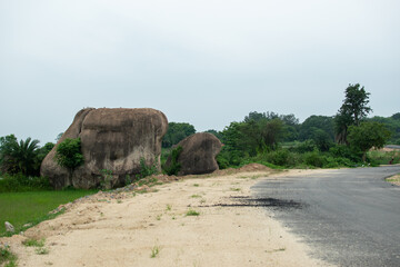 Massive boulders rest beside a winding rural road, surrounded by lush greenery under a calm, cloudy...