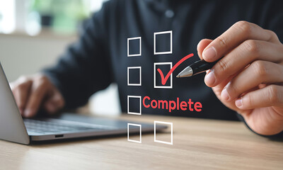 Person checking off a task on a digital checklist using a pen near a laptop on a wooden desk