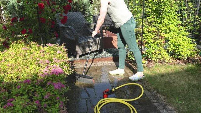 Woman clean outdoor tiles in garden with brush in summer. Buildings in backyard. Cleanliness and order.
