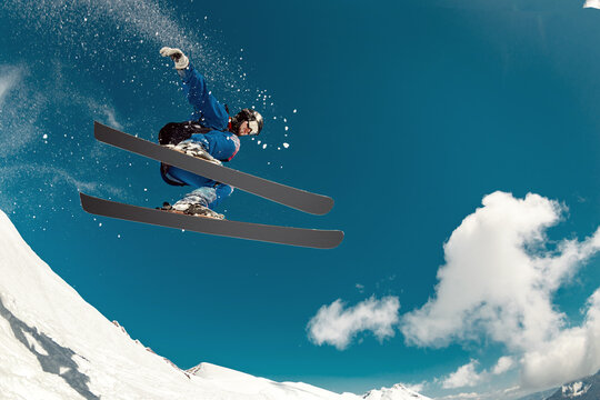 A shot from below of a skier performing a high extreme jump while off-piste skiing against a snowy slope and clear blue sky. Dynamic flight, adrenaline and freedom of freeride
