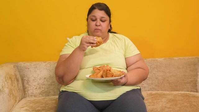 Hungary overweight fat indian woman eating unhealthy samosa junk food. Plus size lady eating fried snack while sitting at home.. Obesity and weight gain. Obese female craving high calorie meal. 4k